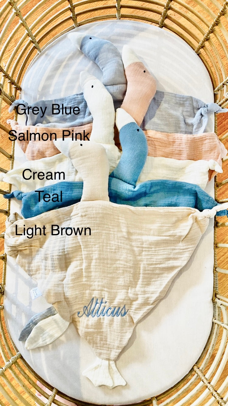 Personalised bird baby comforters stacked in a cot. Grey Blue, Salmon Pink, Cream, Teal and Light Brown bird baby comforters. Top comforter is personalised with Atticus in light blue thread. By Bespoke Munchkin