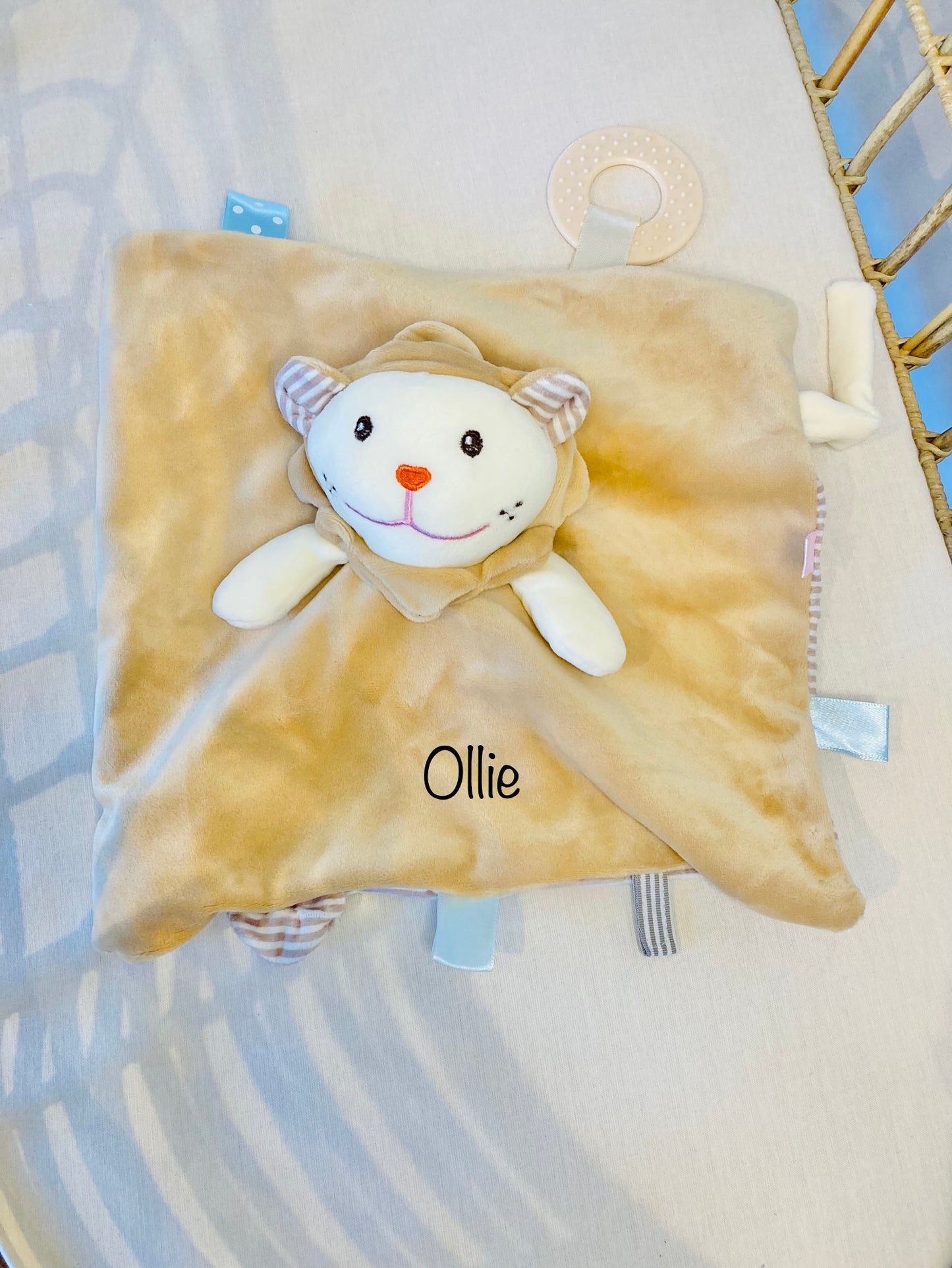 Personalized brown lion baby comforter with the name 'Ollie' embroidered on it, with an attached teether. Comforter is in a cot.