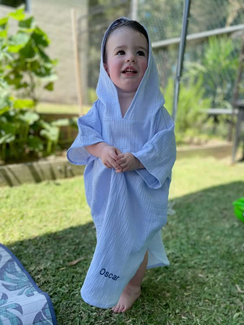 A child standing outdoors wearing a blue hooded towel poncho with a visible embroidered name 'Oscar' at the bottom of the poncho.
