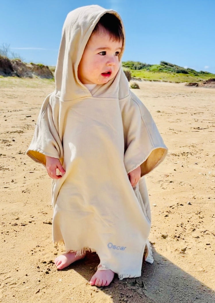 Child on a sandy beach, wearing a personalised cream poncho with name Oscar embroidered at the botton.