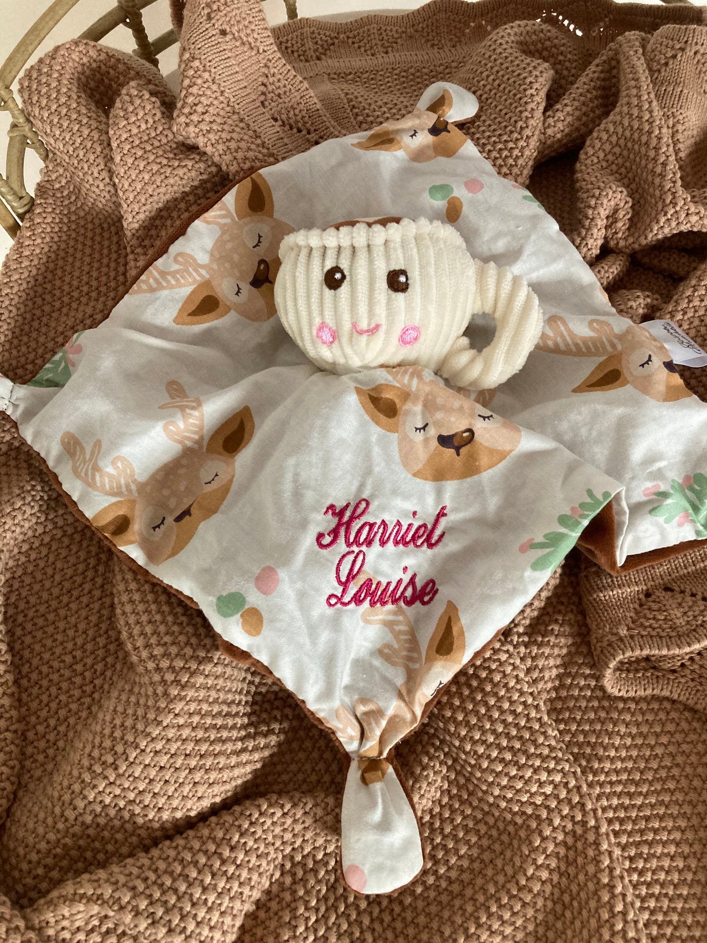 Teacup baby comforter in a cot on top of a baby blanket. Teacup comforter has deer print mini blanket embroidered with baby name attached to a plush toy teacup.