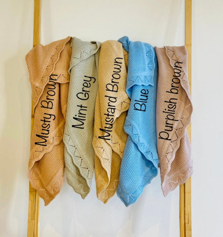 Cotton knitted scalloped edged baby blankets draped over a rack in the following colours from left to right; Musty Brown, Mint Grey, Mustard Brown Blue and Purplish Brown.
