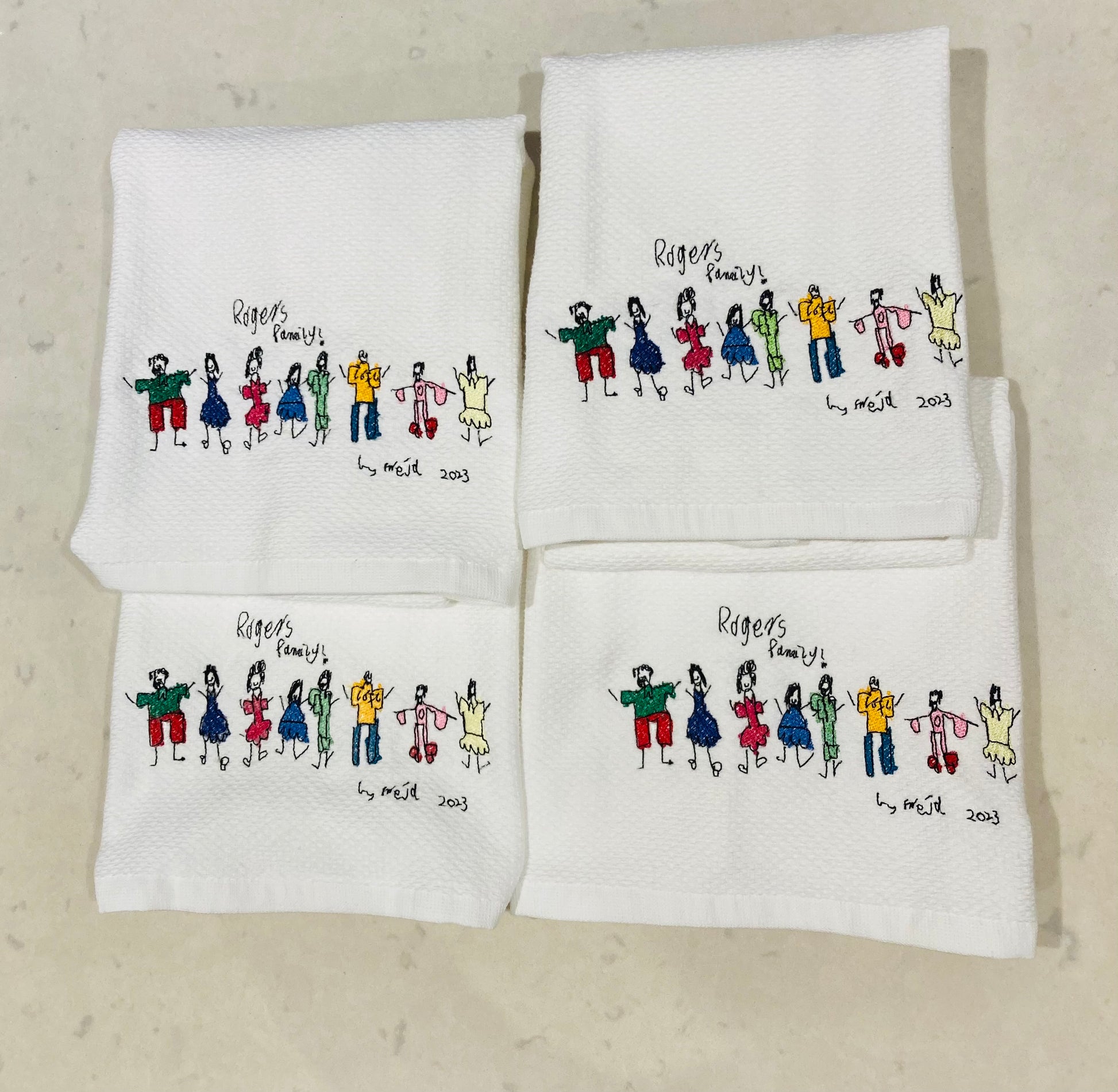 Set of four embroidered towels with colorful family illustrations from a child's drawing and writing, on a kitchen countertop.