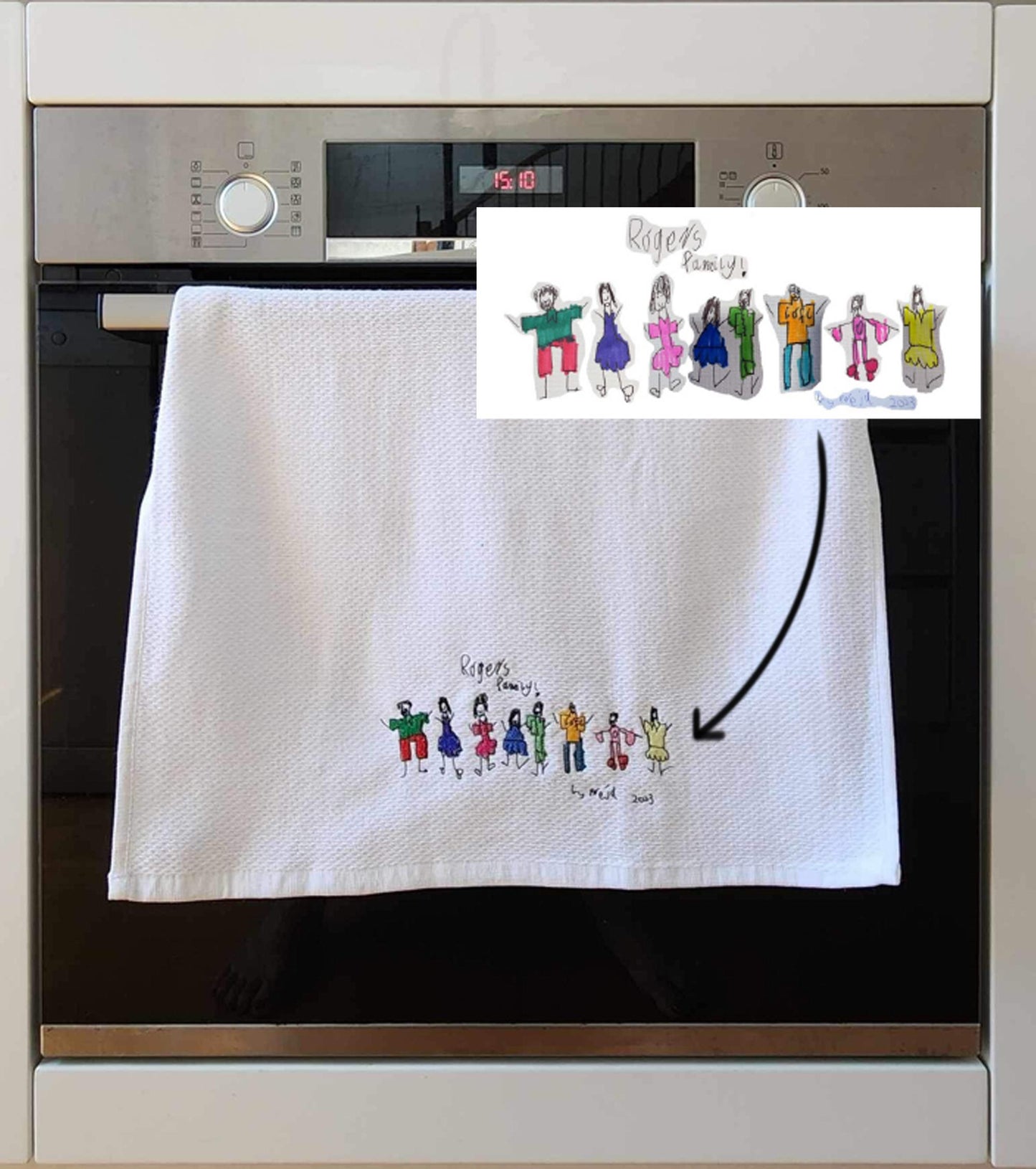White towel with embroidered design of child's artwork, hanging on a kitchen oven.