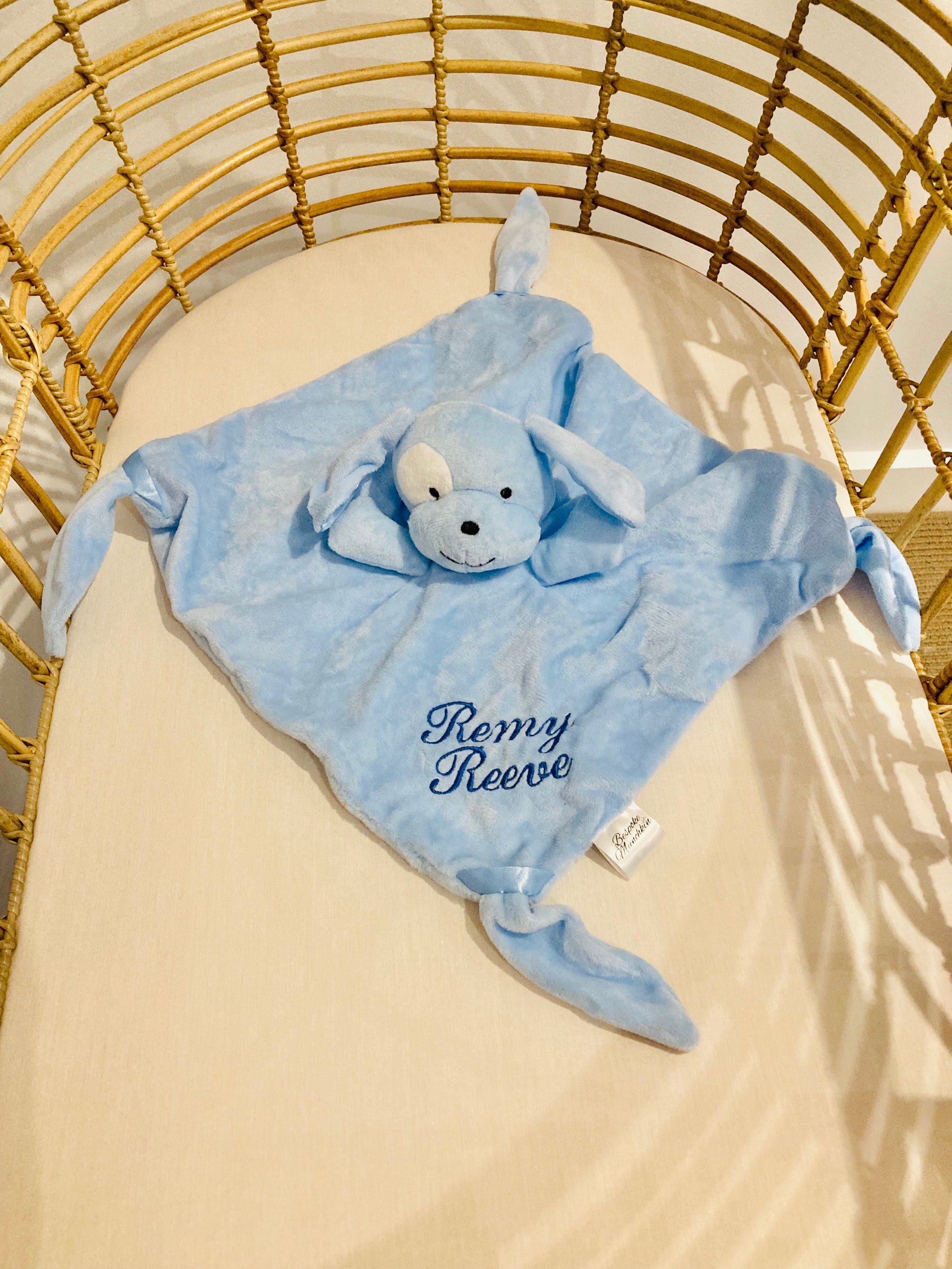 Blue dog baby comforter personalised with an embroidered name Remy Reeve in blue thread. Comforter is in a cot. 