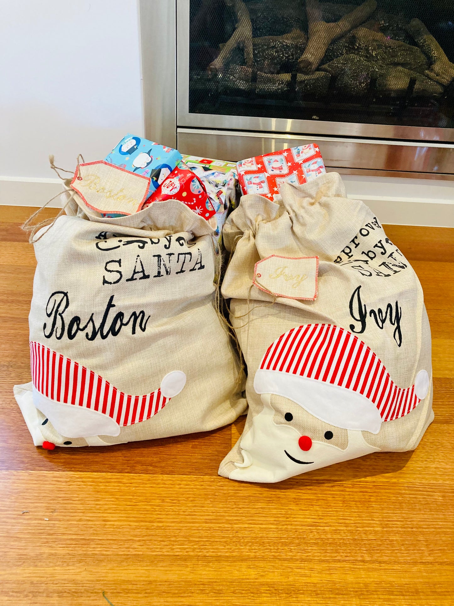 Two Christmas-themed gift sacks with names embroidered on them, one labeled 'Boston' and the other 'Ivy', sitting on a wooden floor in front of a log fireplace.