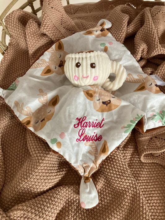Teacup baby comforter in a cot on top of a baby blanket. Teacup comforter has deer print mini blanket embroidered with baby name attached to a plush toy teacup. 