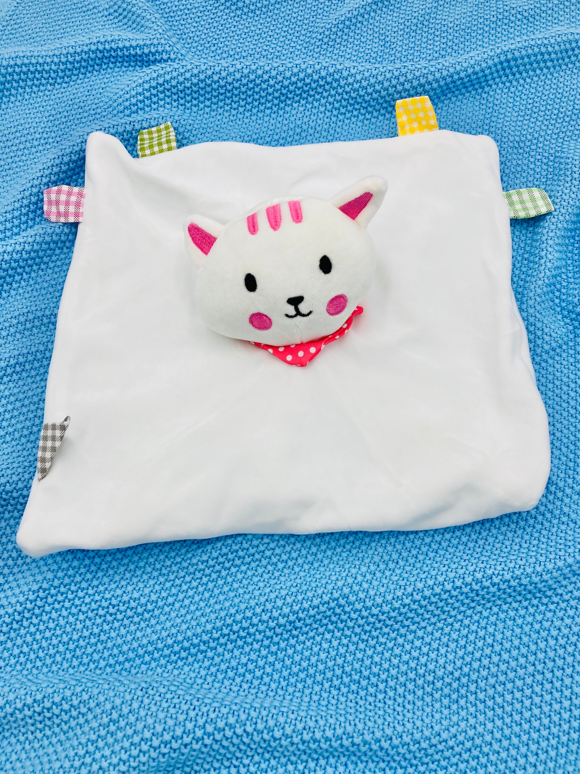A white baby comforter shaped like a cat with embroidered details, colorful tabs, and a rattle, placed on a blue baby blanket.