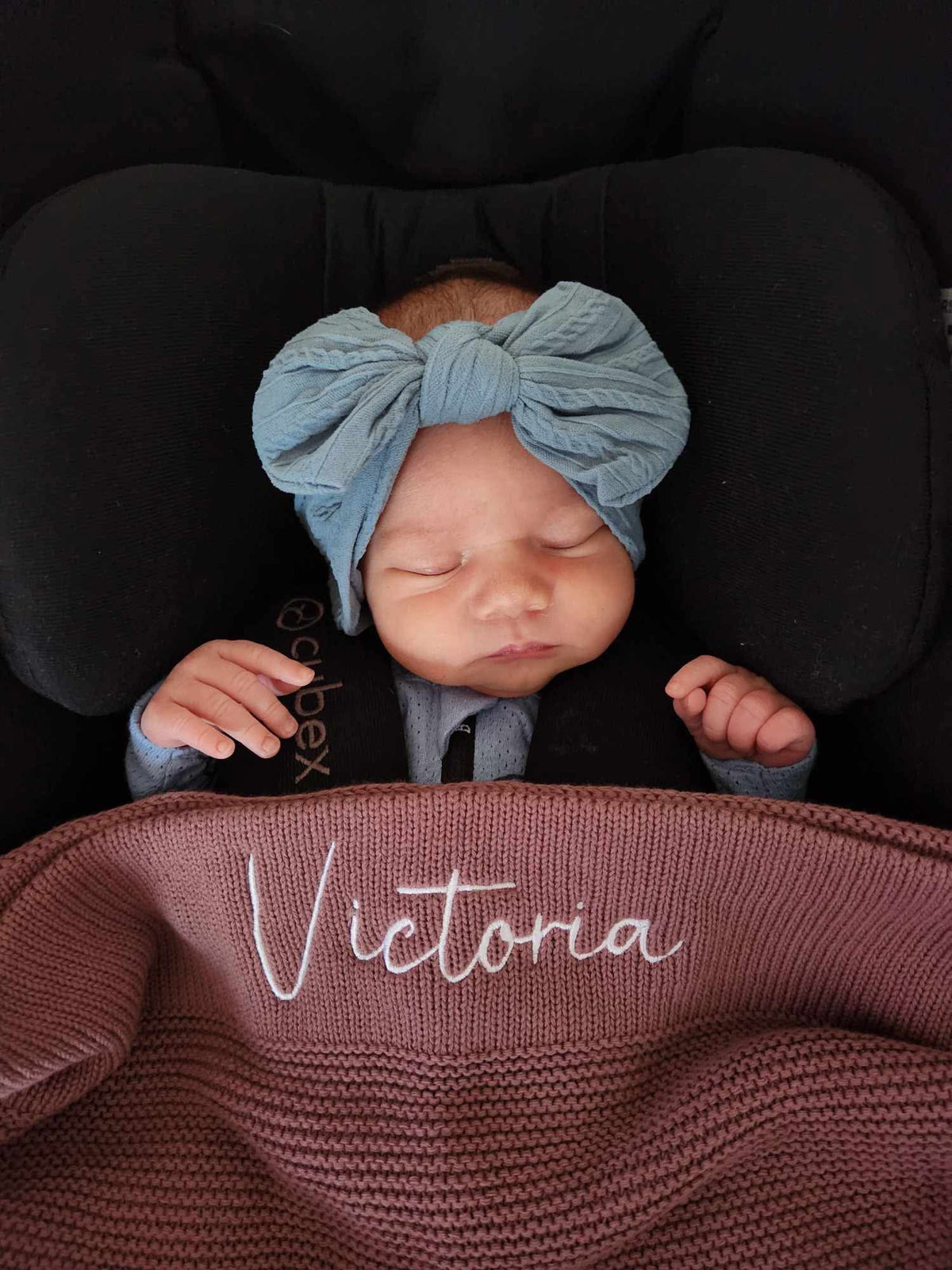 A sleeping newborn baby wearing a large dusty blue bow headband and a dark outfit, resting in a black car seat. A mauve knit blanket is draped over the baby, featuring the name 'Victoria' embroidered in white script.