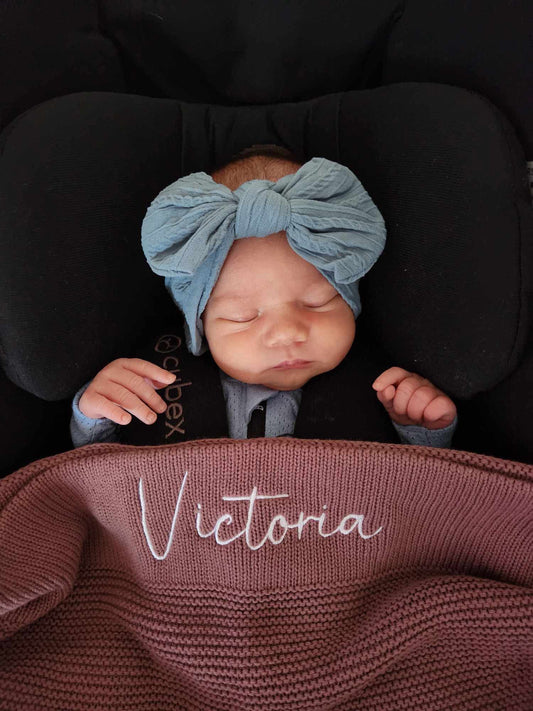 A sleeping newborn baby wearing a large dusty blue bow headband and a dark outfit, resting in a black car seat. A mauve knit blanket is draped over the baby, featuring the name 'Victoria' embroidered in white script.