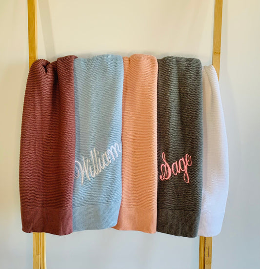 Personalised cotton knit baby blanket with broad rim drapped over a rack. Baby blanket colours from left to right are Ruby Red, Baby Blue, Salmon Pink, Grey and White. The baby blue blanket is embroidered with 'William' in white and the grey baby blanket is embroidered with 'Sage' in light pink.  