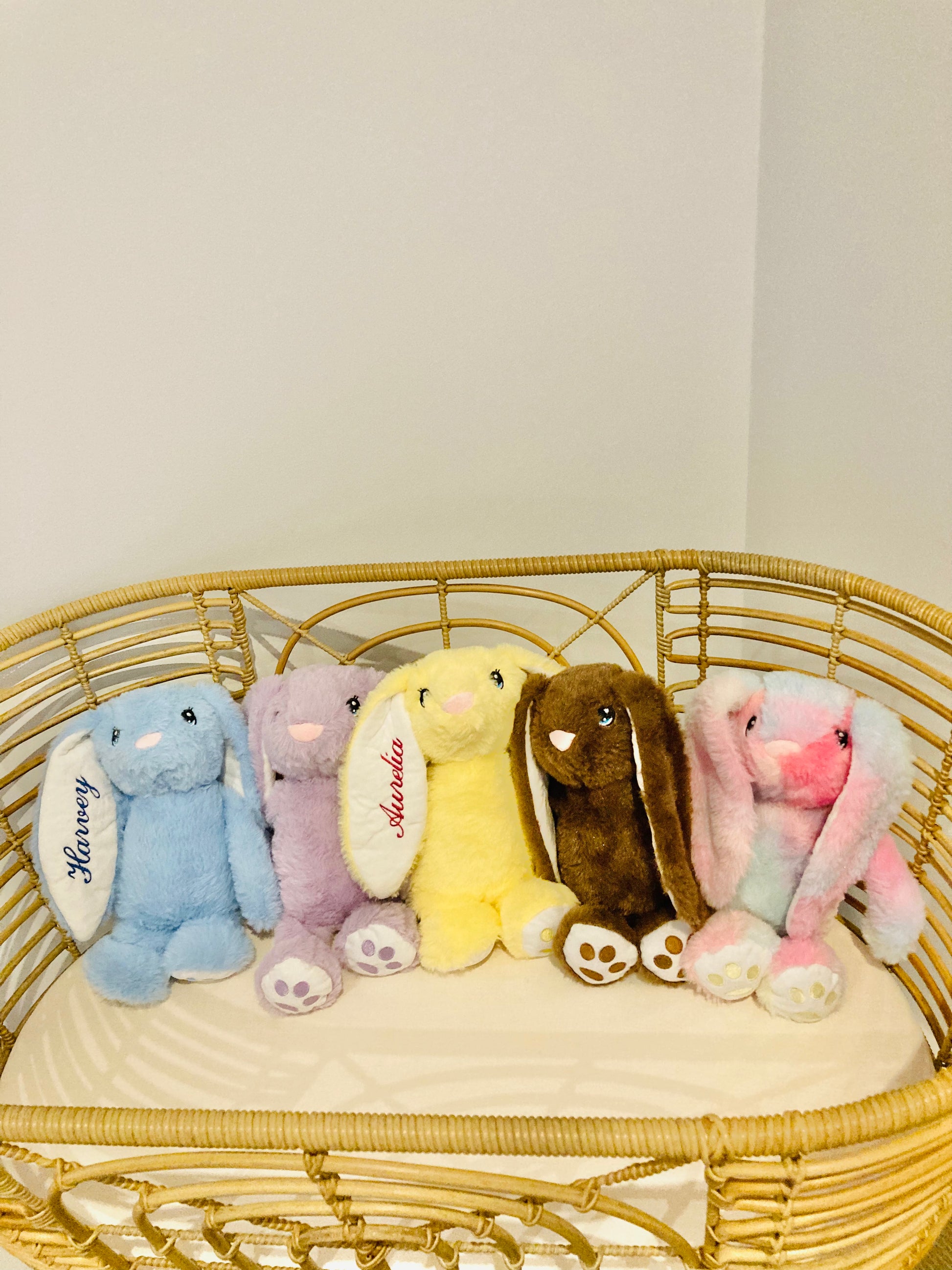 Blue, purple, yellow, brown and rainbow plush bunnies in a cot. The Blue bunny is personalised with Harvey and the yellow bunny is personalised with Aurelia on the ear. 