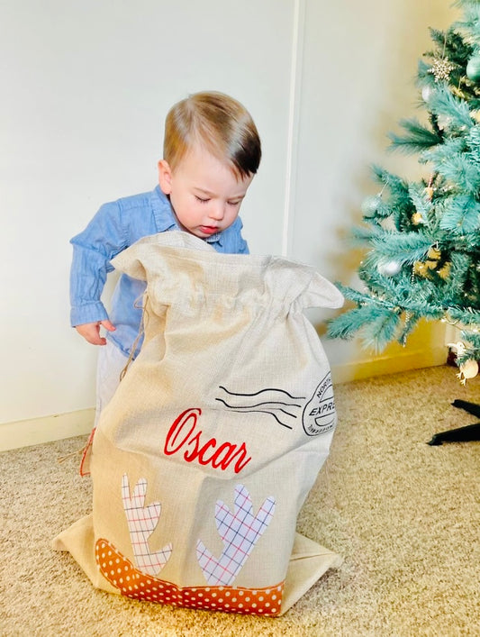 Cute boy looking into his santa sack. Santa has an applique of a reindeer and is embroidered with name 'Oscar' with a Christmas tree in the background. 