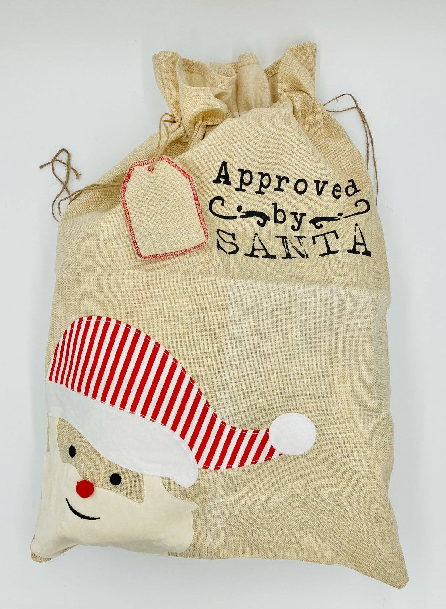 A beige cotton Santa sack with a Christmas design, including a Santa Claus face and a removable gift tag with 'Approved by Santa' text.